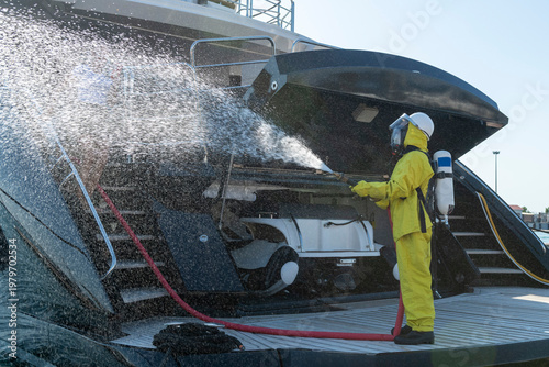 Fire drill on yacht stern: crew member in protective suit uses fire hose and breathing apparatus during training, simulating emergency firefighting on board.
