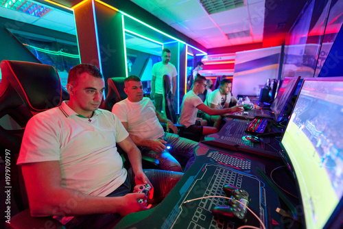 Fotografie Young Men Gaming Together in Neon-Lit LAN Center Playing Console and PC Multipla