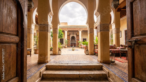 Sunlit Entrance Gate To Tiled Courtyard With Painted Columns And Patterned Steps In Restored North African Guesthouse, Wide Architectural Travel View