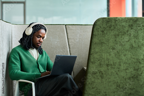 Schilderij op canvas African American young man wearing headphones, working on laptop in office booth, focusing on remote tasks