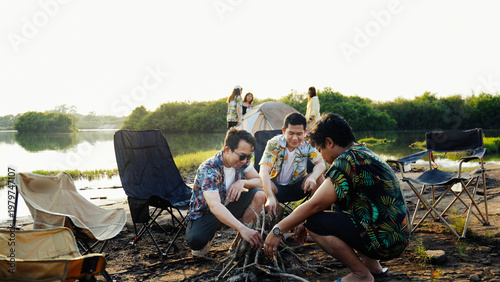 Friends building a wooden campfire together near their lakeside tent, enjoying warm sunlight and relaxed summertime camping moments outdoors.