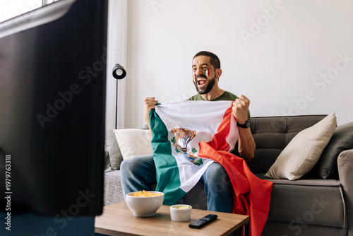 Mexican football fan celebrating at home while holding national flag and watching a match on TV. Adult man cheering on the sofa with snacks and drinks in the living room.