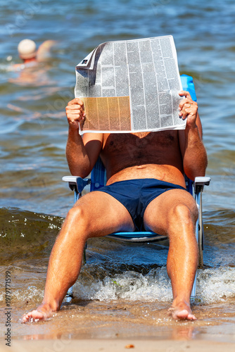 A man is relaxing in an armchair right by the waves, engrossed in his reading and covering his face with an open newspaper