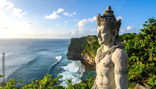 Carved stone figure overlooks cliffside ocean, Bali. Sky with clouds in background, tropical foliage