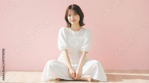 Young Japanese woman sitting cross-legged in a white dress on pink background