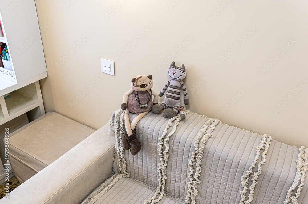 Naklejka premium Two plush cat toys sit on a light grey quilted sofa with fringed details. A white shelving unit and ottoman are nearby; a light switch is visible on the beige wall behind.