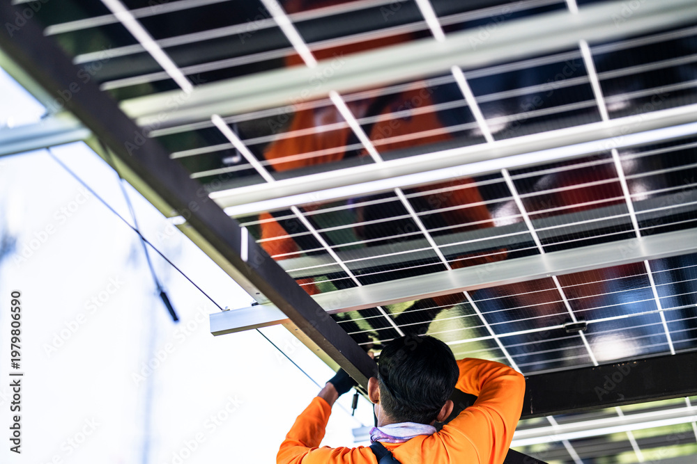 Fototapeta premium A technician in an orange safety jacket working on the wiring and connections of a solar panel system installed on a steel carport structure. The worker is focused on maintenance or installation.