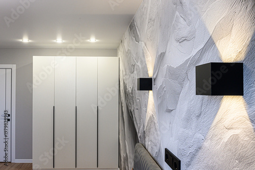 A modern interior with a textured rock wall, sleek white wardrobe, recessed ceiling lights, and two black square wall lamps casting soft light on the decorative surface.
