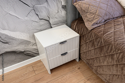 A white bedside тумбочка with two drawers and sleek black handles stands next to a quilted brown bed, against a textured grey wall, on a wooden floor.