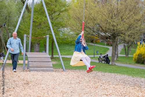Grandfather watching granddaughter on zip line, happy young girl playing, smiling man, playground family fun, outdoor play, active childhood, nature movement and joy, healthy lifestyle family time