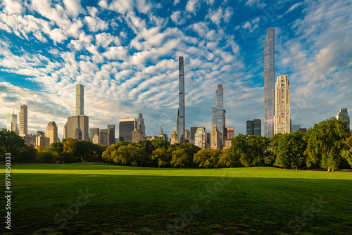 Lush green grass and trees provide a natural foreground to the skyline of Midtown Manhattan from Central Park with the tall and thin ultra luxury skyscrapers of Billionaires Row in New York City.