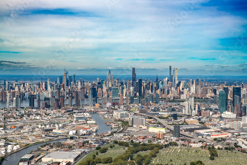 A wide aerial view shows the dense urban landscape of Queens in the foreground with the iconic Midtown Manhattan skyline across the East River in New York City.