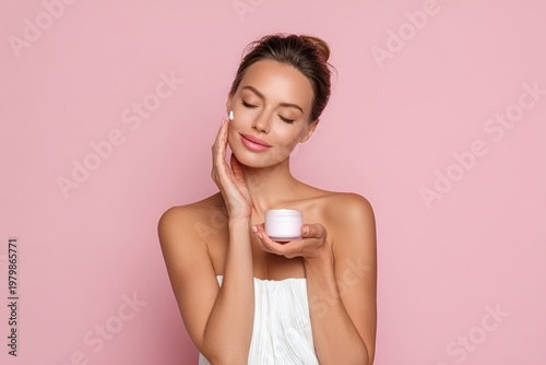 Woman applying cream to her face while holding a small jar in front of a pink background during a skincare routine