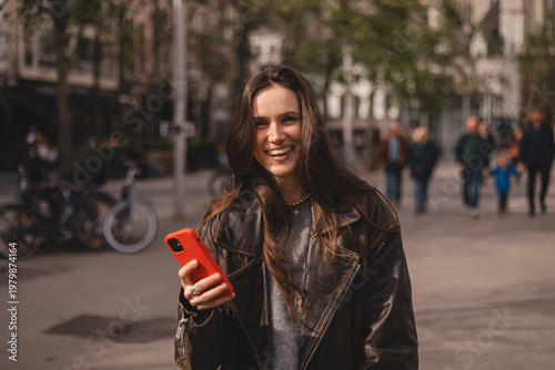 Cheerful young woman in leather jacket laughing while using red smartphone on busy city street, happy emotional portrait in urban environment, digital communication and lifestyle concept.