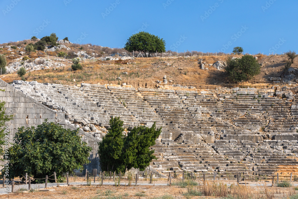 Obraz premium Ruined Roman Theatre of Patara ancient city. A historic stone amphitheater ruins with terraced seating, an arched entrance, and weathered walls sits in a sunlit landscape. Turkey