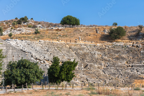Ruined Roman Theatre of Patara ancient city. A historic stone amphitheater ruins with terraced seating, an arched entrance, and weathered walls sits in a sunlit landscape. Turkey