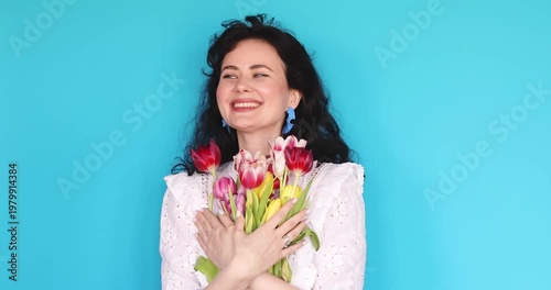 Happy Young Caucasian Woman with Curly Hair Enjoying and Embracing a Gift Bouquet of Colorful Tulips, Showing Delightful Facial Expressions, Closing Eyes in Pleasure on Blue Studio Background