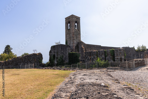 Medieval castle ruins with tall bell tower in Cavriana Lombardy Italy, wide ground level view follows rough stone path past dry grass and fenced remains toward weathered walls rising