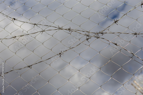 Close-up of a chain-link fence topped with barbed wire against a blurred sky background.