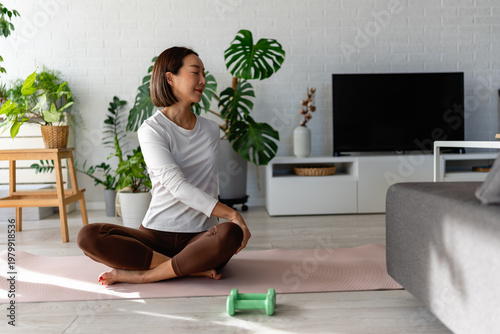 Photos East Asian woman sitting cross-legged on a yoga mat, gently twisting her torso during a home workout