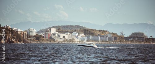 Telephoto coastal cityscape with speedboat crossing choppy bay, waterfront buildings, wooded hill and hazy mountain range under soft daylight, scenic urban shoreline panorama