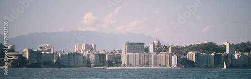Telephoto panorama of coastal city skyline across bay water, with dense waterfront towers, sandy shoreline, distant mountain range, soft haze, pastel sky and scenic urban coast