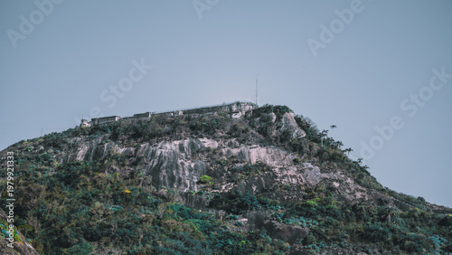 Rocky hillside with historic fort on the summit, weathered stone slopes, sparse tropical vegetation and wide blue sky, scenic elevated landmark in natural mountain landscape