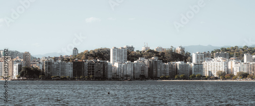 Telephoto view of waterfront skyline across calm bay, with dense residential towers, wooded hills, faint mountain range and wide pale sky, creating a scenic urban coastal panorama