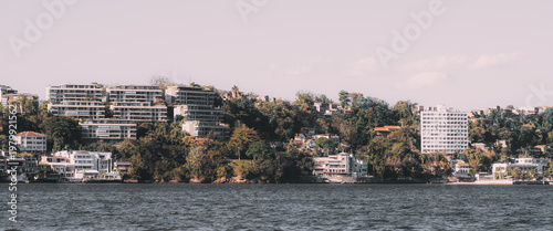 Telephoto view of upscale waterfront neighborhood with modern apartment buildings, lush hillside villas, calm bay water and soft pastel sky, capturing a scenic residential coastal cityscape