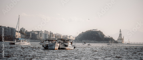 Telephoto coastal bay scene with fishing boats and catamaran on choppy water, backed by hazy city shoreline, rocky island, distant bridge and offshore platform under soft pastel sky
