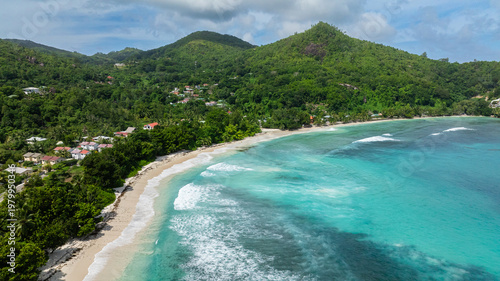 Clear turquoise waves wash over large smooth rocks on the white sandy shore, surrounded by greenery. Seychelles, Mahe. Baie Lazare.