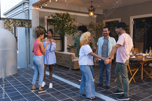 Diverse friends chatting, holding hands on patio with string lights, wicker sofa, wood table