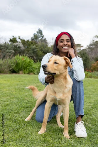 Fototapeta Adult Asian woman kneeling on lawn at park wearing red headband holding golden dog collar, smiling