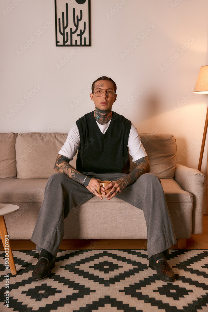 Fototapeta premium Handsome young man dressed in stylish attire sits on a sofa in a warm, cozy room