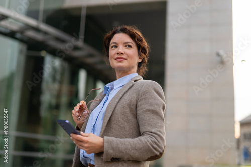 Фотография Confident businesswoman holding smartphone and glasses looking ahead