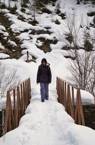 A young girl hiking with a backpack, walking across a snow-covered bridge. 