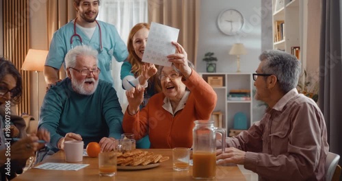 Excited elderly woman winning bingo during group game in assisted living home. Happy winner celebrating success, joy and congratulations with other seniors indoors.