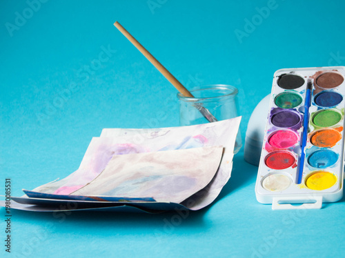 Colorful watercolor paint in plastic palettes, paintbrush in glass and paper sheets of watercolor children's drawings. Creative mess, blue table background