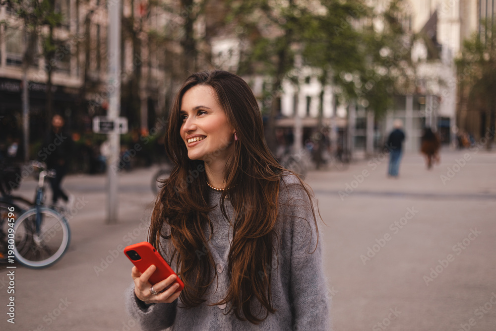Fototapeta premium Dreamy young woman with long brunette hair smiling and looking away while holding red smartphone on blurred city street background, charming girl in grey cardigan enjoying morning in the city