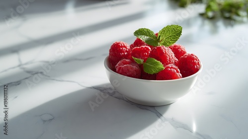 A bowl of fresh raspberries with mint leaves on a marble countertop in sunlight