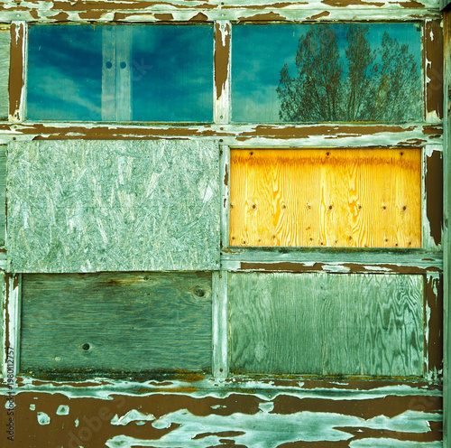 Old wooden garage door has patches & old glass with reflection. Panels are boarded up. Building looks like junk. Door framework has chipped curling paint so bare wood is visible.