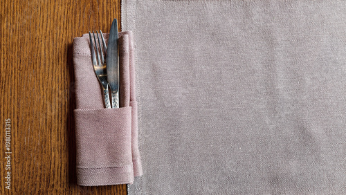 An overhead shot of a fork and knife placed in a light pink napkin, resting on a wooden table next to a patterned placemat. The composition is simple and elegant with warm lighting