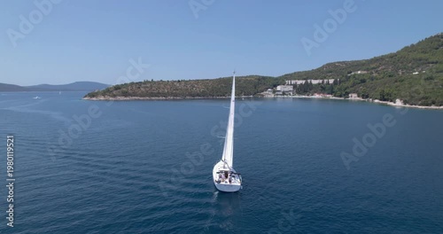 Sailboat Entering Sheltered Bay Near Rocky Headland And Low Coastal Development, Glassy Water Shows Subtle Wake And Reflections, Drone Tracks Steady Approach