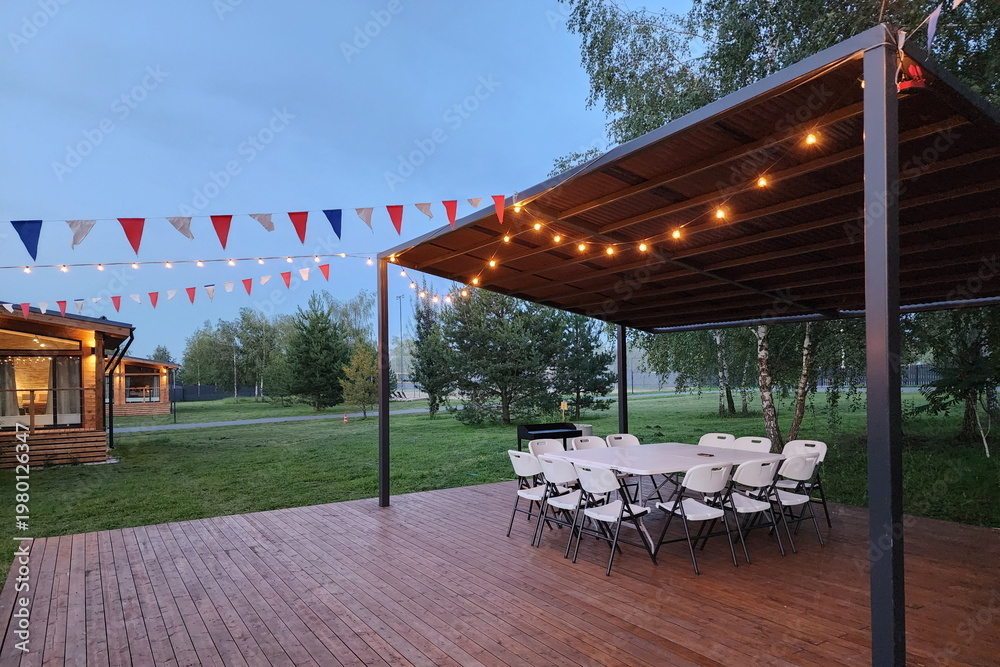 Naklejka premium A gathering area features folding chairs set in a circle under a wooden structure. Strings of lights hang above colorful decorations. The scene takes place in an open green space at dusk
