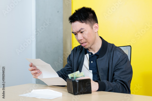 An Asian male student frowns while holding a bill in one hand and checking his wallet with cash at a desk, ideal for student budget struggles, financial stress, or cost of living concepts.