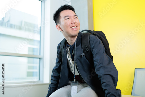 An Asian male student wearing a backpack and ID lanyard laughs while looking upward in a bright campus room, ideal for student orientation, university life, or international student welcome concepts.