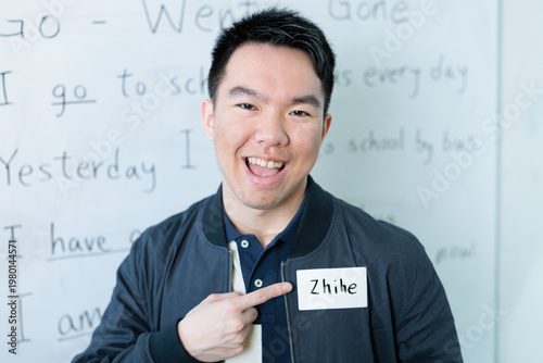 An Asian male student grins while pointing to his name tag in front of a whiteboard with English grammar sentences, for student introduction, ESL or international student orientation concepts.