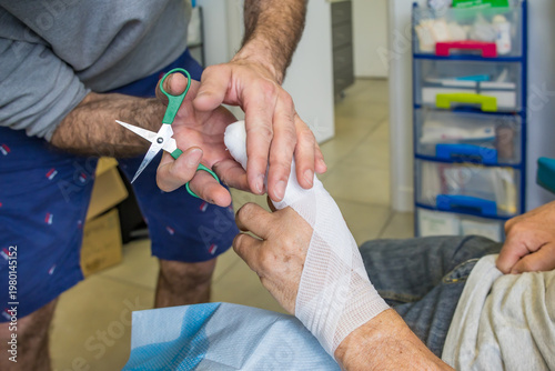 Tableau sur toile Nurse Cutting Bandage with Scissors While Wrapping Elderly Patient Hand in Outpa