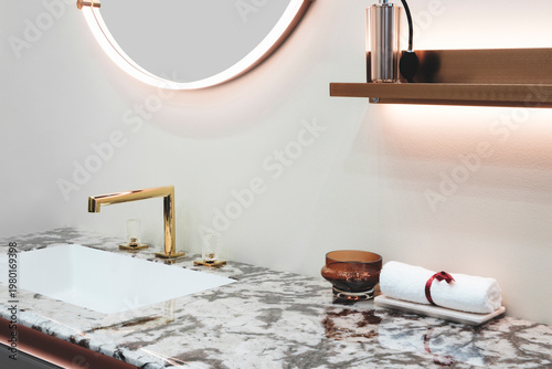 Modern bathroom vanity with white vessel sink and gold faucet. Wall-mounted mirror glows softly above. Towel hangs neatly on brass holder