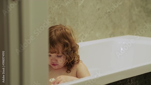 Little girl with curly hair sitting in white bathtub. Cheerful child enjoying bath time in bathroom. Concept of hygiene, childhood care, happy home life and daily routine for youngster.